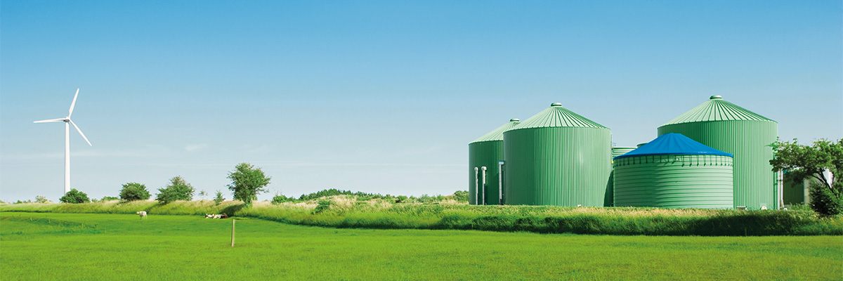 Vista de instalaciones con depósitos verdes cilíndricos y un aerogenerador en un campo abierto con césped y cielo despejado