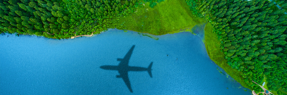 Sombra de un avión proyectada sobre un lago azul rodeado de un paisaje verde con árboles y campos